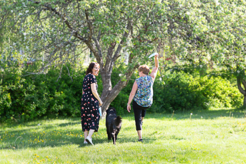 två personer och en hund går i lummig sommarmiljö